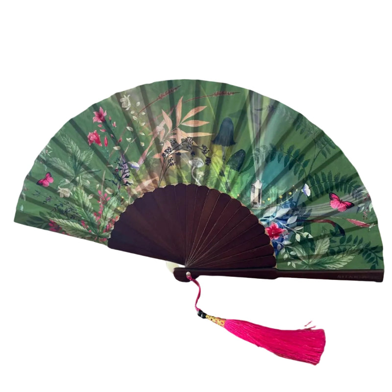 Floral-patterned hand fan with a pink tassel on a wooden surface, with flowers and a plant in the background.