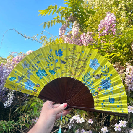 Hand holding a yellow floral fan with a blue tassel against a backdrop of trees and flowers.