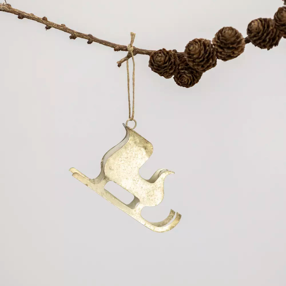 A gold-toned metal Christmas ornament shaped like a sleigh with a bird silhouette, hanging from a twig garland with pinecones against a light gray background.