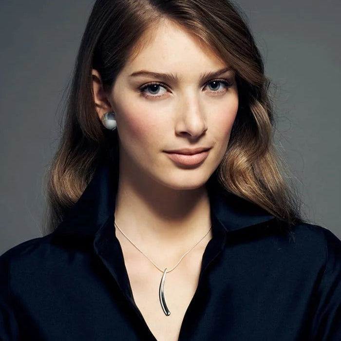 A woman with brown hair wearing a black collared shirt, a silver pendant necklace, and a silver stud earring, photographed against a dark gray studio background.