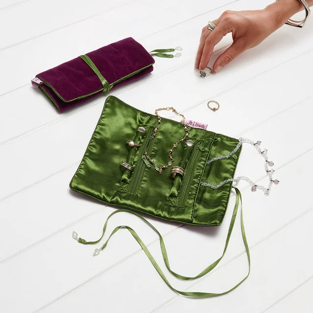 A close-up of jewelry storage pouches on a white surface, with a hand reaching toward a small silver ring holder. The pouches are made of shiny green satin and deep purple velvet, both decorated with small gold-tone rings.