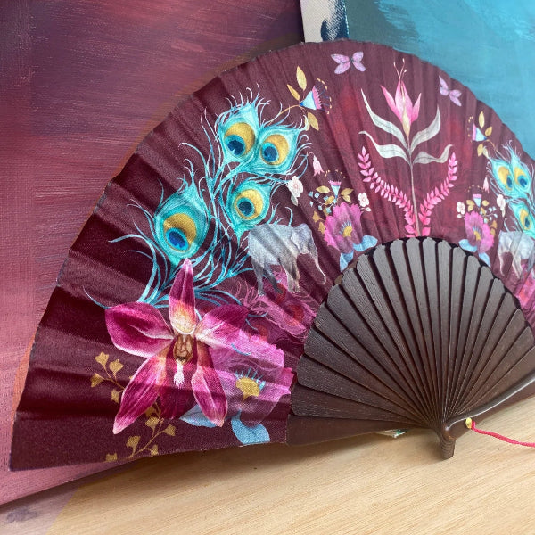 A folding hand fan with dark brown wooden ribs and a burgundy fabric panel decorated with colorful floral and peacock feather motifs, resting on a light wood surface.
