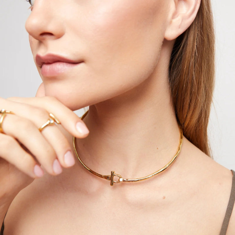 A close-up lifestyle shot of a model wearing a delicate gold bangle choker necklace with a T-shaped clasp, along with gold rings.