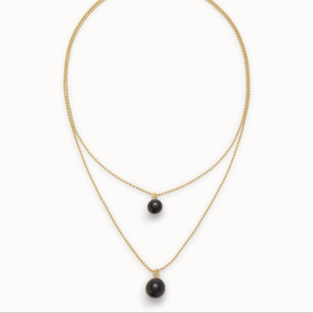 A gold double-strand necklace featuring a small round black bead pendant on the upper chain and a larger black bead pendant on the lower chain, displayed against a clean white background.