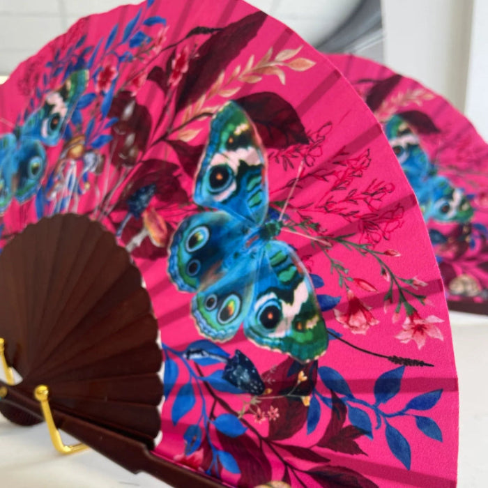 A folding hand fan with a bright pink floral background and a large blue-and-green butterfly print, attached to a dark brown wooden handle with gold-tone hardware. A second matching fan is partially visible behind it on a light surface.