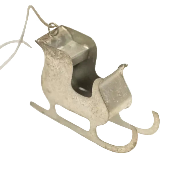 A distressed silver metal sleigh ornament with a hanging loop, shown on a clean white background with a soft-focus green plant in the corner.
