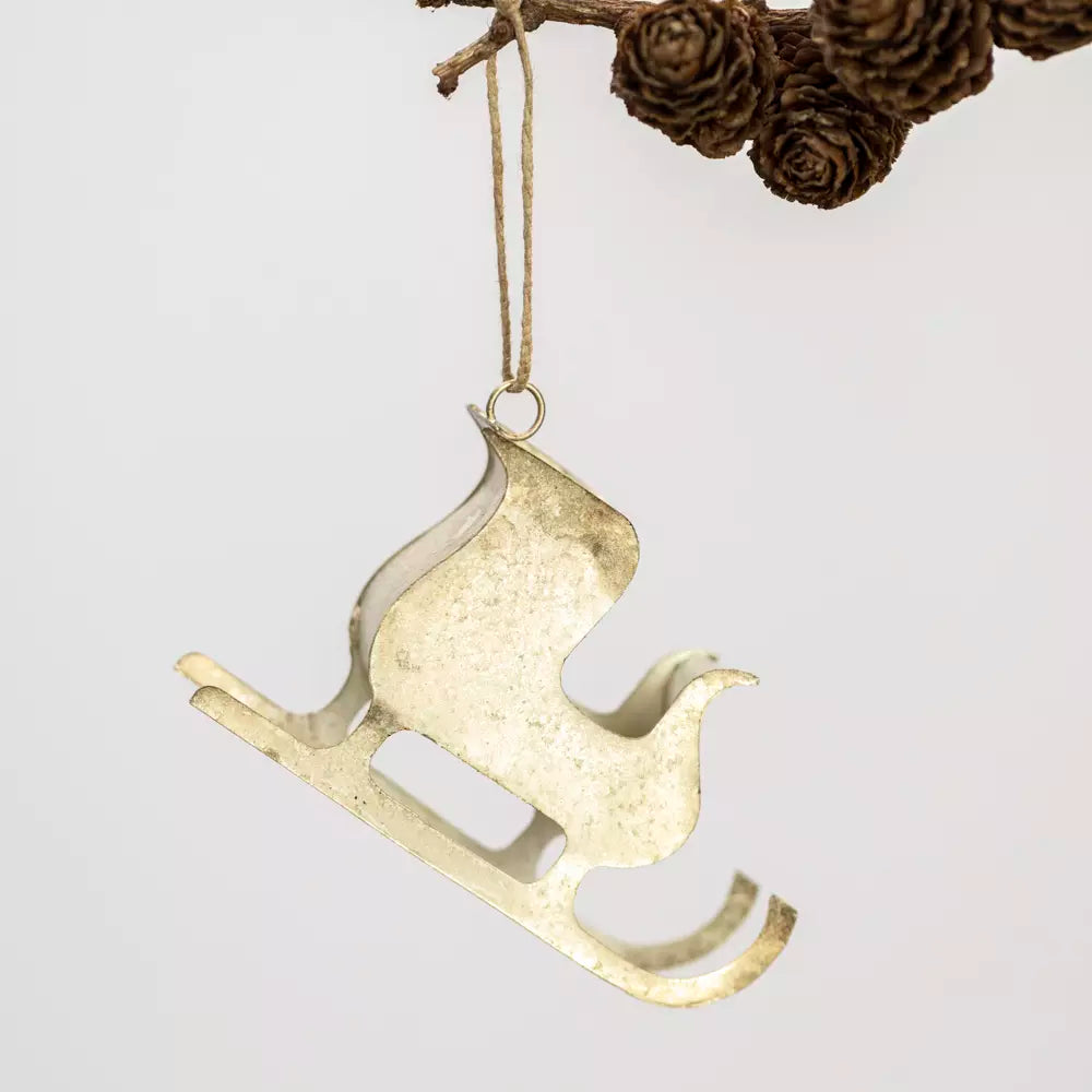 A distressed gold metal Christmas ornament shaped like a sled, hanging from a jute string attached to a pine branch with pinecones against a light gray background.