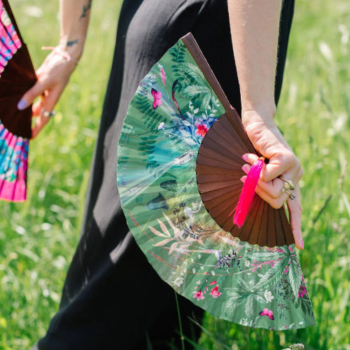 A person outdoors in a grassy field holding a folding hand fan with a green tropical floral print and a dark brown wooden frame with a bright pink tassel.