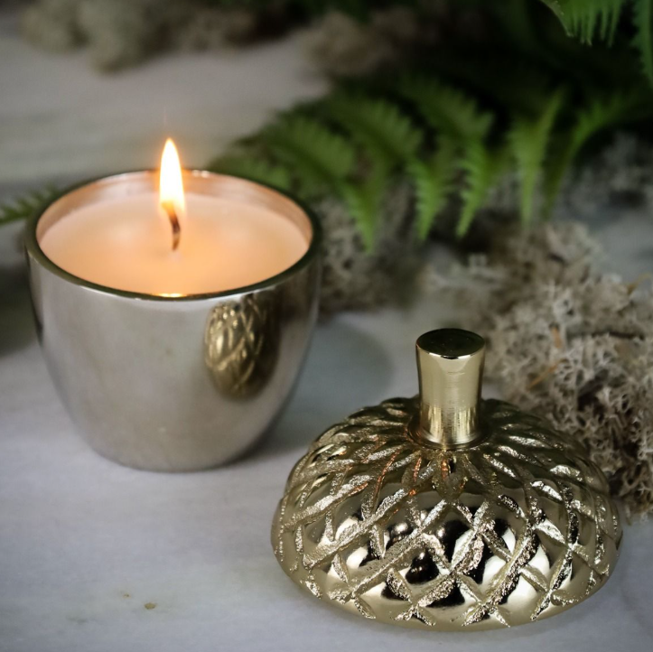 A lit silver candle holder with a flame beside a decorative gold lid with a textured lattice pattern, styled on a light surface with green ferns in the background.