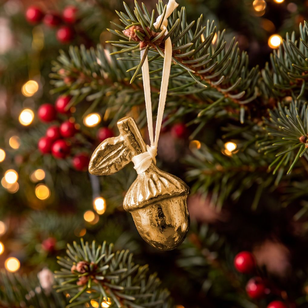 A metallic gold acorn Christmas ornament hanging from a green pine tree branch with red berries and warm bokeh string lights in the background.