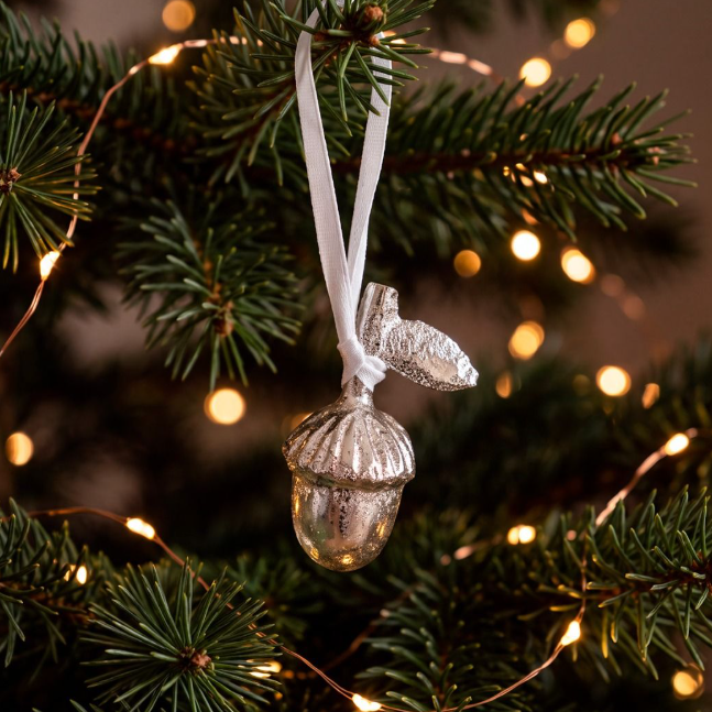 A silver acorn Christmas ornament with a white ribbon hanger, hanging from a green pine tree branch with warm bokeh string lights in the background.