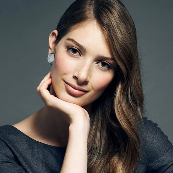 A woman with long brown hair posing in a dark gray top, wearing a thin silver bangle bracelet on her wrist, set against a solid dark gray studio background.