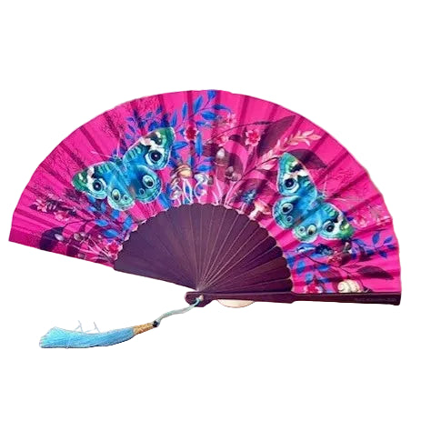A folding hand fan with a bright pink background and detailed blue butterfly illustrations, displayed on a wooden table with blurred flowers and a potted plant in the background.