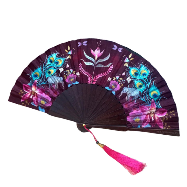 A folding hand fan with a dark purple background and colorful floral and peacock feather designs, featuring a bright pink tassel, displayed on a light wood surface against a pink and blue painted backdrop.